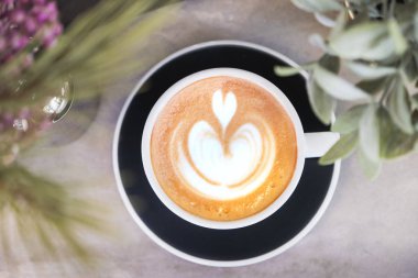 Yeşil bitki ve zemin ön plan, yiyecek ve içecek konsepti ile gri beton sayaçta sıcak cappuccino kahve latte sanat ile siyah Kupası'nda üstten görünüm
