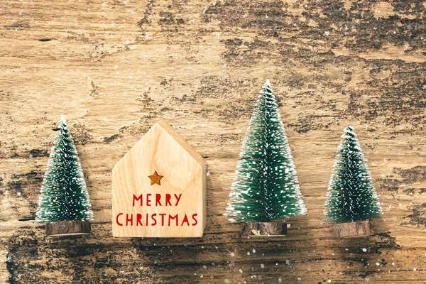 Top view of handwriting red Merry Christmas on home toy with mini christmas tree wood on rustic wooden table with snow fall.winter holiday seasonal greeting card.leave space to adding text or design