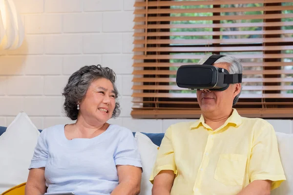 Asian senior couple play virtual realtiy glasses headset and tablet watching vr video and  have fun together on sofa  in living room at home.senior with technology.aging at home