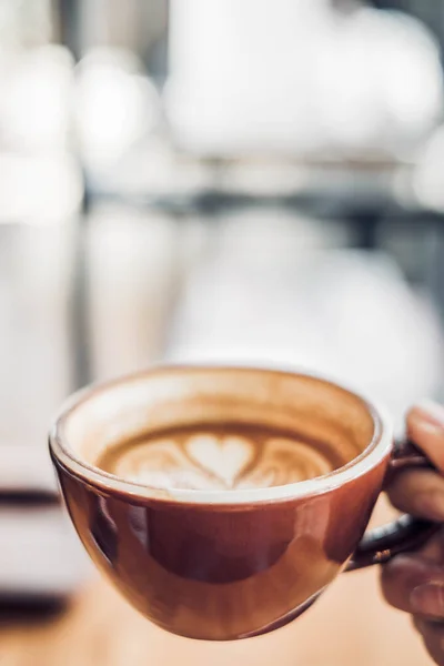 Cerrar la mano sosteniendo una taza de café capuchino caliente taza con forma de corazón latte arte desenfoque café interior.food y bebida concept.copy espacio para añadir texto — Foto de Stock