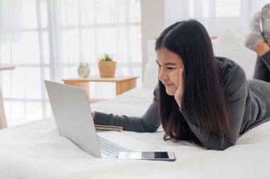 Asyalı kadın yatak odasında dizüstü bilgisayarda evrak yapıyor yatakta yatıyordu.w
