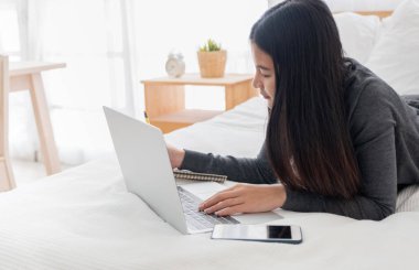 Asyalı kadın yatak odasında dizüstü bilgisayarda evrak yapıyor yatakta yatıyordu.w