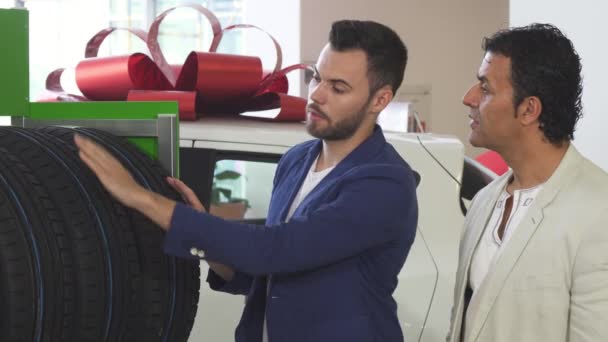 Молодой продавец помогает своему зрелому клиенту выбирать шины для автомобилей — стоковое видео