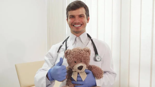 Feliz doctor mostrando el pulgar hacia arriba sosteniendo un oso de peluche —  Fotos de Stock