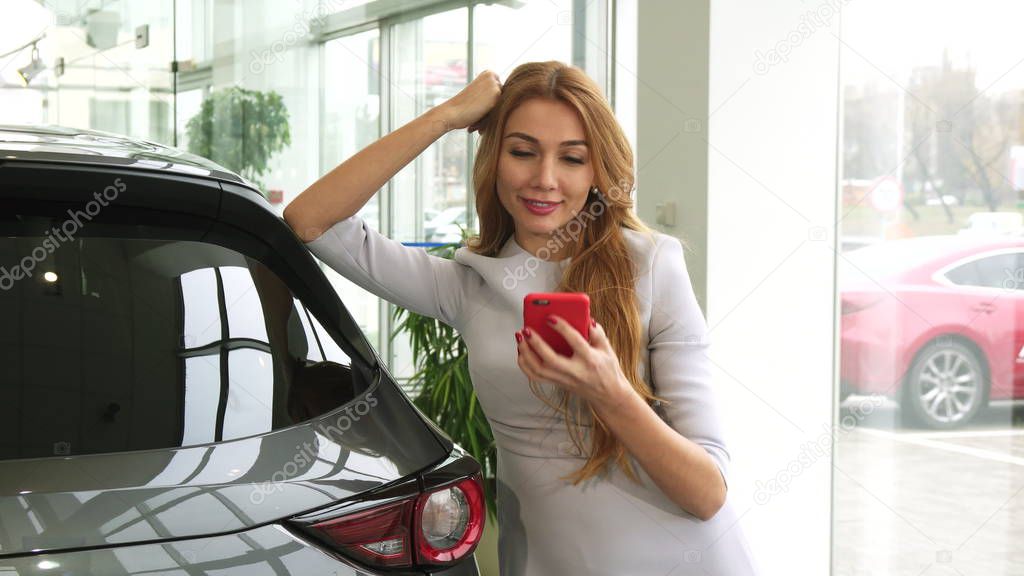 Happy beautiful woman using her smart phone at the car dealership salon