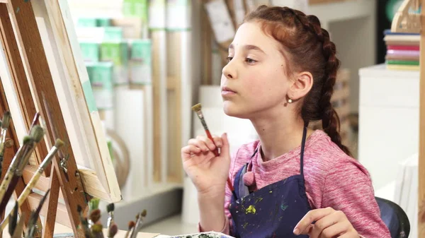 Linda niña mostrando pulgares hacia arriba mientras que la pintura de un cuadro en la clase de arte — Foto de Stock