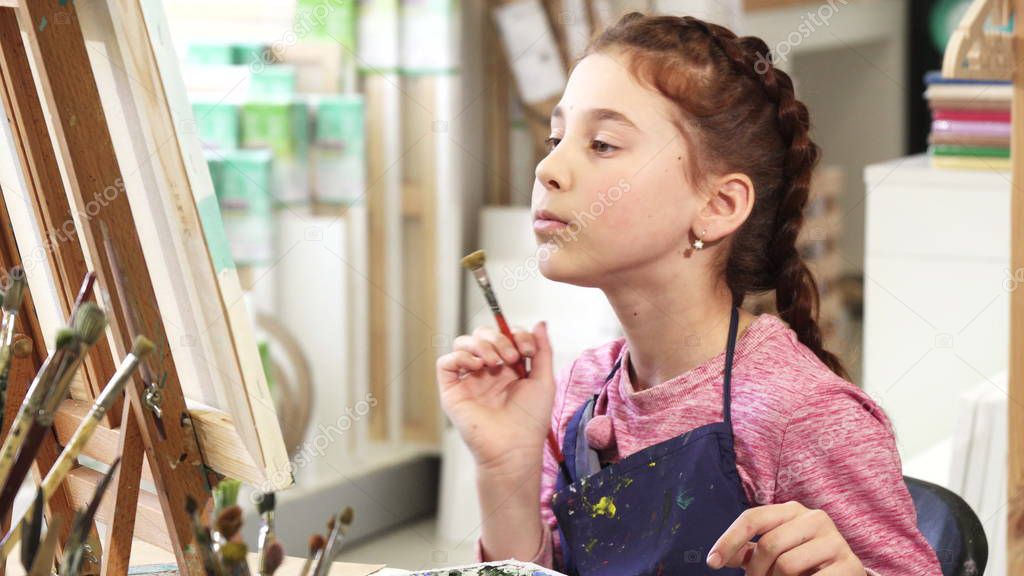 Cute little girl showing thumbs up while painting a picture at the art class