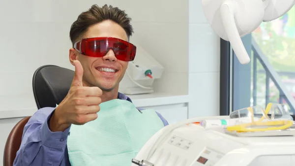 Αρσενικός ασθενής φορώντας προστατευτικά γυαλιά δείχνει τους αντίχειρες στο οδοντιατρείο — Φωτογραφία Αρχείου