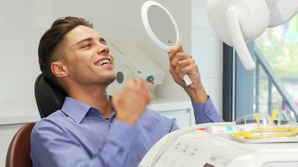 Handsome young man examining his teeth in the mirror at the dental clinic — Stock Photo, Image