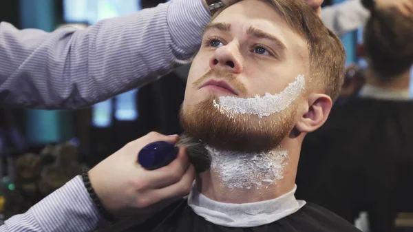 Парикмахер наносит крем для бритья на бороду своего клиента — стоковое фото