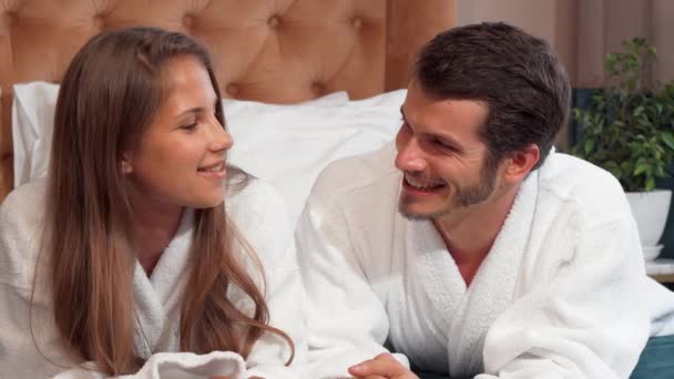 Mutlu sevgi dolu çift birlikte bornoz giyiyor yatakta yatan konuşuyor — Stok video