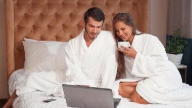 Genç çift yatakta dizüstü bilgisayar kullanırken kahve. Muhteşem bir kadın kocasıyla yatakta kahve yudumlarken gülüyor. Yakışıklı neşeli adam ve karısı dizüstü birlikte otel odasında kullanma