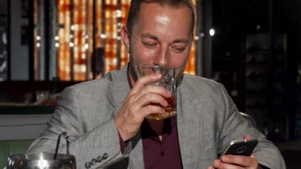 Красивый зрелый мужчина пользуется смартфоном, пробуя виски в баре — стоковое видео