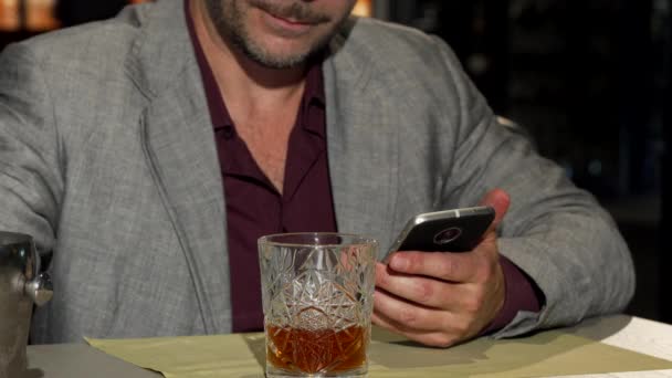 Hombre maduro usando un teléfono inteligente mientras agrega cubitos de hielo a su whisky — Vídeo de stock