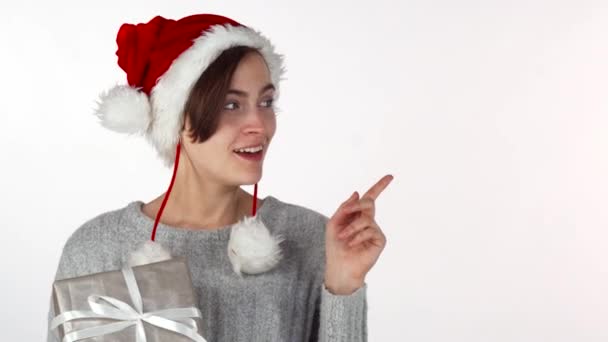Щаслива красива жінка в різдвяному капелюсі тримає подарунок, вказуючи на копійку — стокове відео