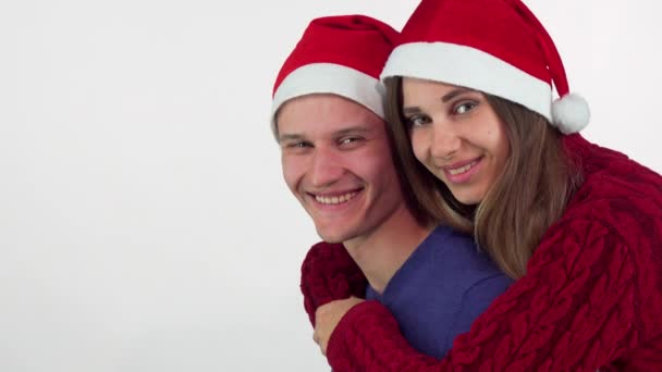 Счастливая молодая пара в рождественских шляпах, обнимается, весело смотрит в сторону — стоковое видео
