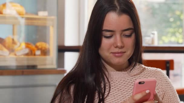 Mujer joven sonriendo, usando su teléfono inteligente en la cafetería — Vídeos de Stock