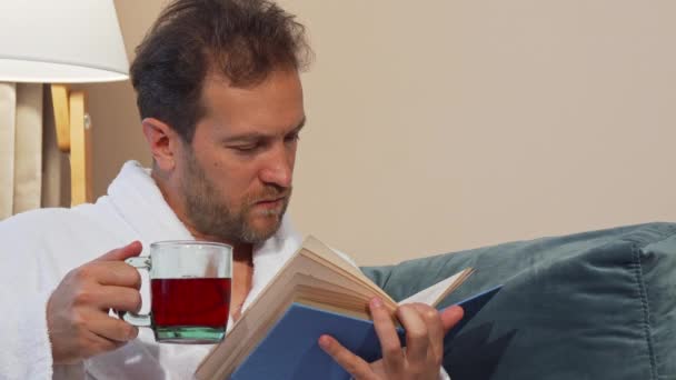 Зрілий чоловік має ароматний гарячий чай, читає книгу вдома, одягнений в халат — стокове відео