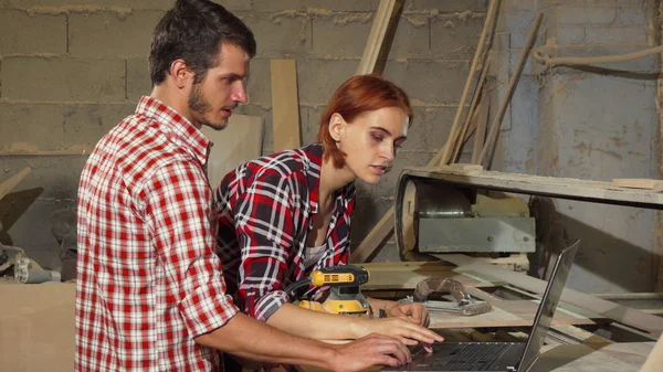 Two carpenters using laptop at their workshop while making furniture