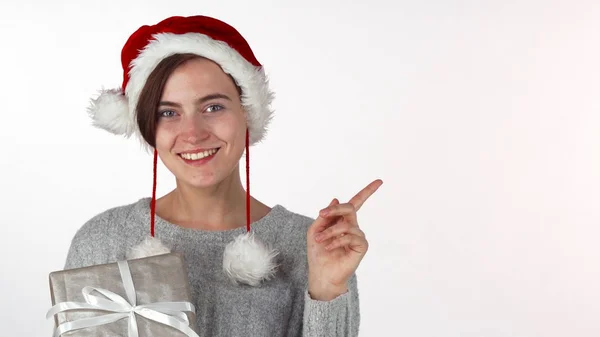 Mulher bonita feliz no chapéu de Natal segurando um presente, apontando para copyspace — Fotografia de Stock