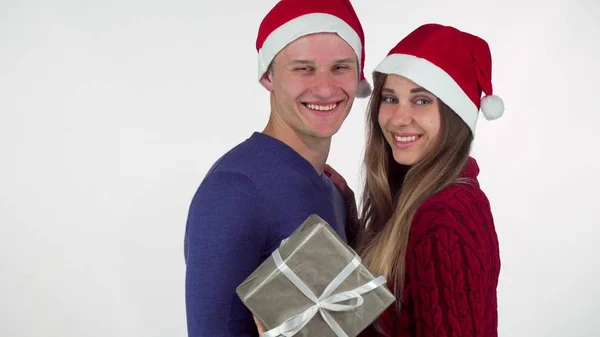 Joven pareja cariñosa con sombreros de Navidad sonriendo a la cámara, sosteniendo un regalo —  Fotos de Stock