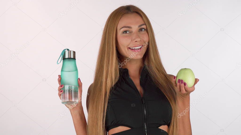 Beautiful sportswoman smiling cheerfully, holding green apple and water bottle