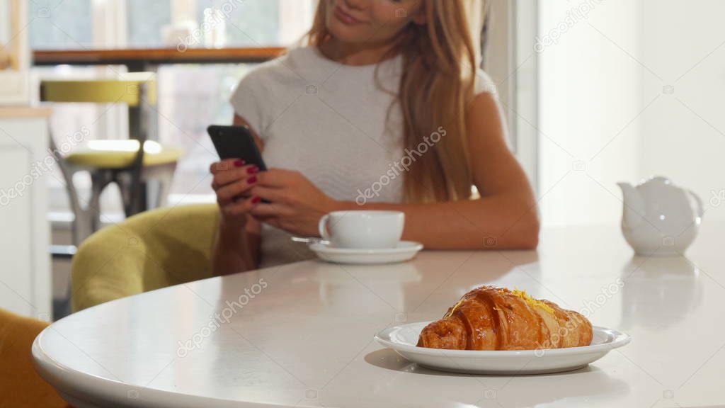 Woman using smart phone at bakery cafe, delicious croissant on the foreground