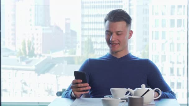 Веселый красивый мужчина радостно улыбается, пользуется смартфоном, пьет чай. — стоковое видео