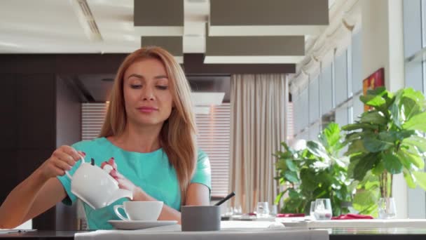 Preciosa mujer feliz vertiendo té en su taza, disfrutando del desayuno en el restaurante — Vídeos de Stock