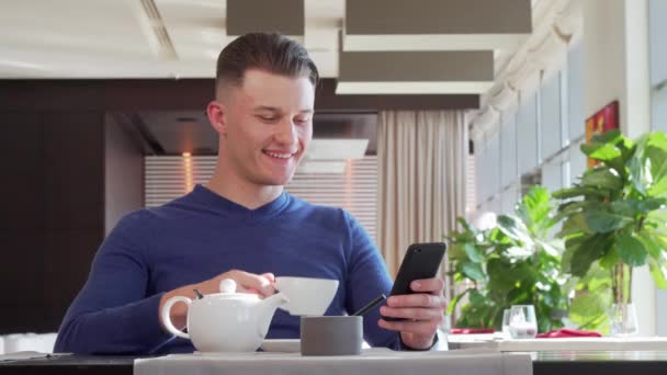 Hermoso hombre feliz riendo, usando su teléfono inteligente en el café durante el desayuno — Vídeos de Stock