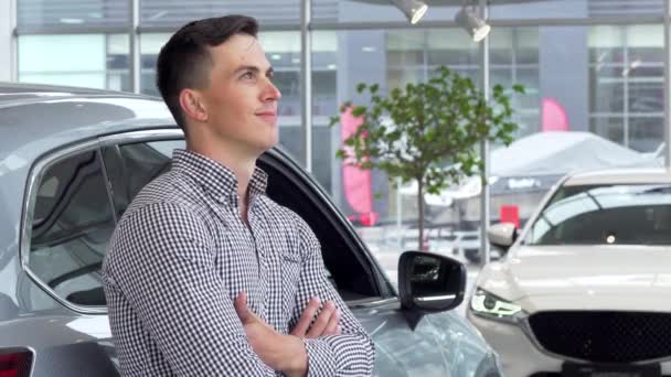 Hombre guapo sonriendo soñando, apoyándose en un coche nuevo en el concesionario — Vídeo de stock