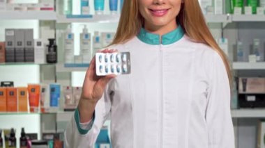 Gülümseyen kadın eczacı blister hap kameraya uzatarak. Doz vitamin paketi tutan eczanede, çalışma mutlu bir kimyager kırpılmış. Perakende, tıp kavramı