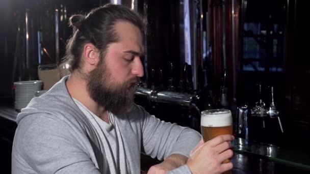 Бородатый длинноволосый мужчина, выглядящий подавленным, пьющий пиво один в баре — стоковое видео
