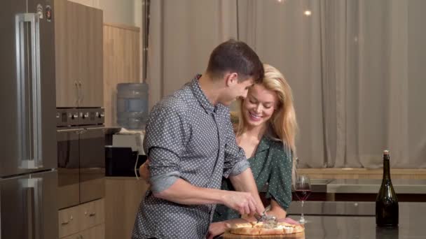 Bonito homem cortando pizza em casa, sua esposa amorosa abraçando-o — Vídeo de Stock