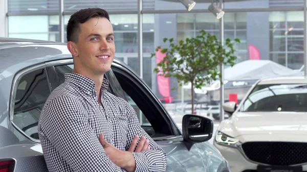 Schöner Mann, der verträumt lächelt und sich im Autohaus an ein neues Auto lehnt — Stockfoto