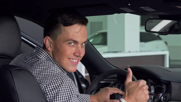 Красивый мужчина улыбается через плечо, показывая большие пальцы вверх, сидя в новой машине — стоковое фото