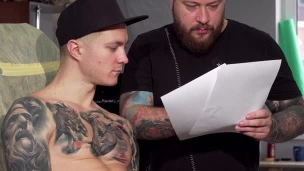 Skäggiga tatuering artist diskuterar skisser med hans manliga klient — Stockvideo