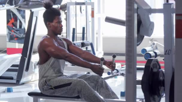 Hombre africano atlético guapo haciendo ejercicio en la máquina de fila baja sentada en el gimnasio — Vídeos de Stock