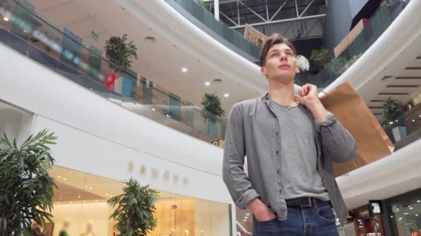 Χαμηλή γωνία ενός νεαρού άντρα που κρατά σακούλες για ψώνια, κοιτάζοντας γύρω από το εμπορικό κέντρο — Αρχείο Βίντεο