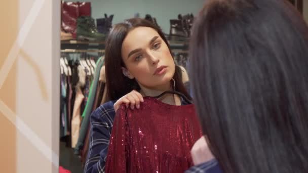 Красивая женщина выбирает между двумя платьями, покупки в магазине одежды — стоковое видео