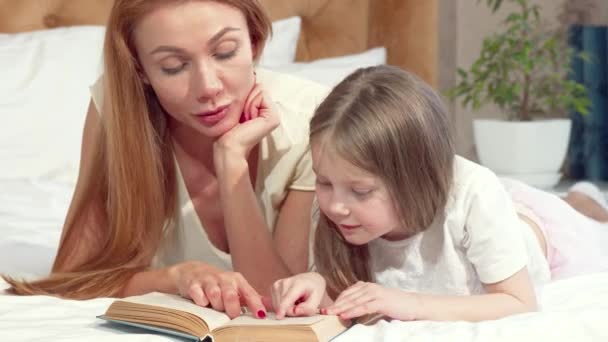 Schöne Frau liest ein Buch mit ihrer niedlichen kleinen Tochter, zu Hause auf dem Bett liegend — Stockvideo