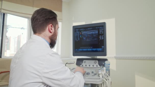 Веселий лікар посміхається до камери, використовуючи машину для ультразвукового сканування — стокове відео
