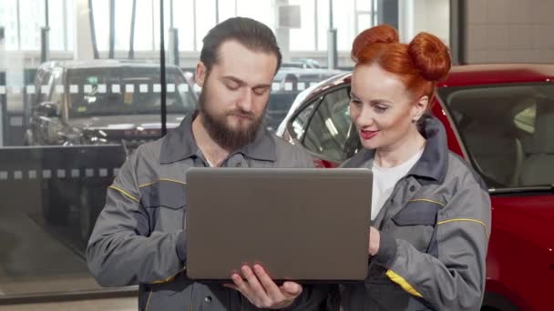Mecánico de coche barbudo usando el ordenador portátil en el garaje, trabajando con una colega — Vídeos de Stock