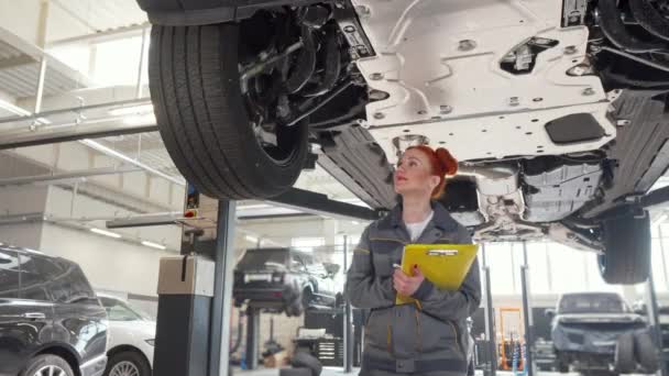 Vrouwelijke automonteur lopen onder de auto op een lift, het schrijven van aantekeningen op het Klembord — Stockvideo