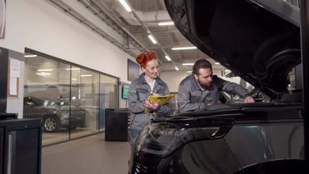 Dos mecánicos profesionales examinando el motor de un coche, tomando notas — Vídeos de Stock