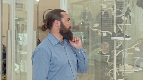 Зрілий бородатий чоловік задумливо дивиться на дисплей магазину одягу — стокове відео