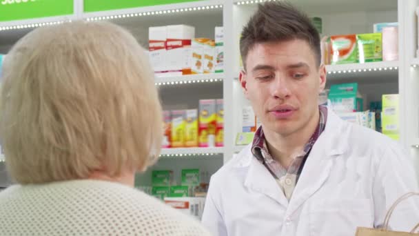 Красивый молодой фармацевт вручает сумку с покупкой клиенту — стоковое видео