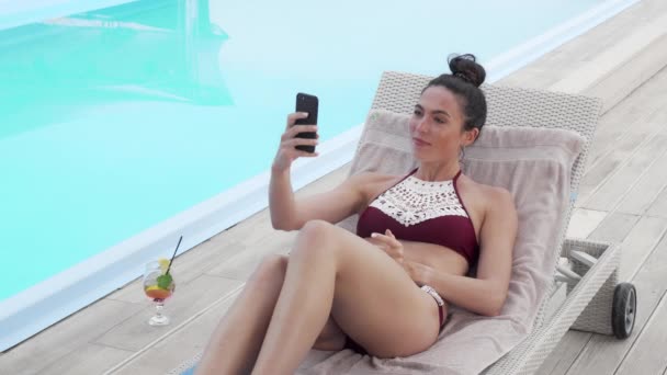 Красивая женщина делает селфи со своим смартфоном, отдыхая у бассейна — стоковое видео