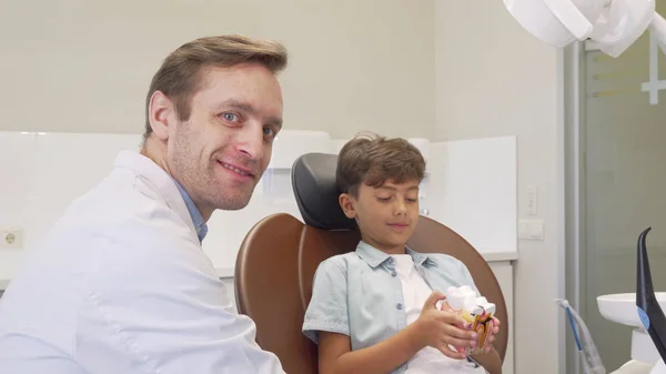 Mogen tandläkare leende till kameran medan du arbetar med bedårande liten pojke — Stockfoto