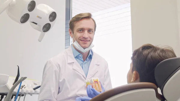 Cheerful mature male dentist smiling to the camera while educating little patient — Stock Photo, Image